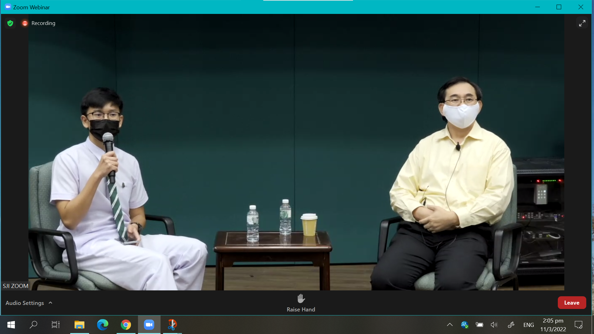 Two masked people sit in chairs during a Zoom Webinar recording, one holding a microphone, with a small table between them.